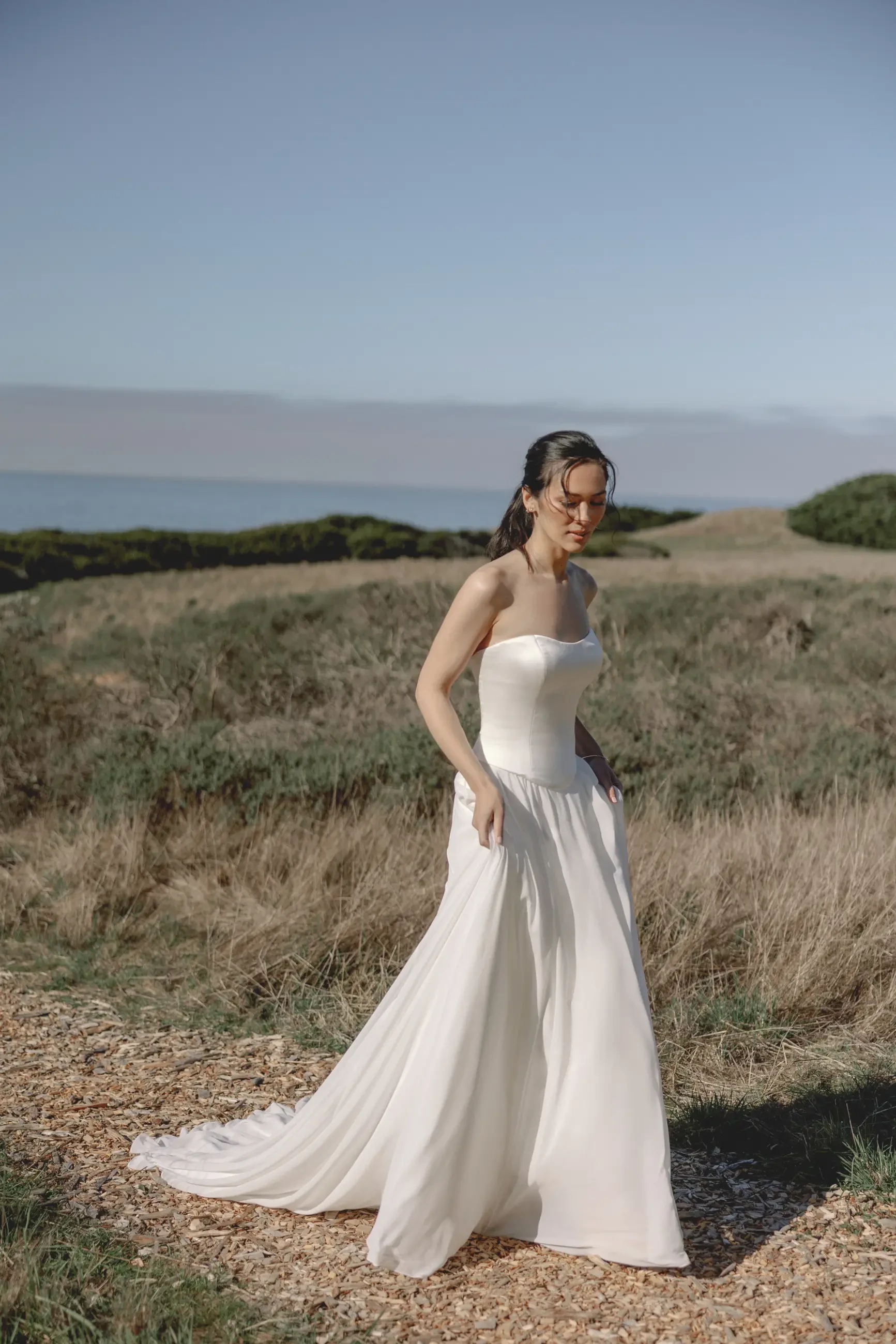 A woman in a white strapless wedding dress stands on a grassy coastal path, with the ocean and clear sky in the background. She appears serene and reflective.