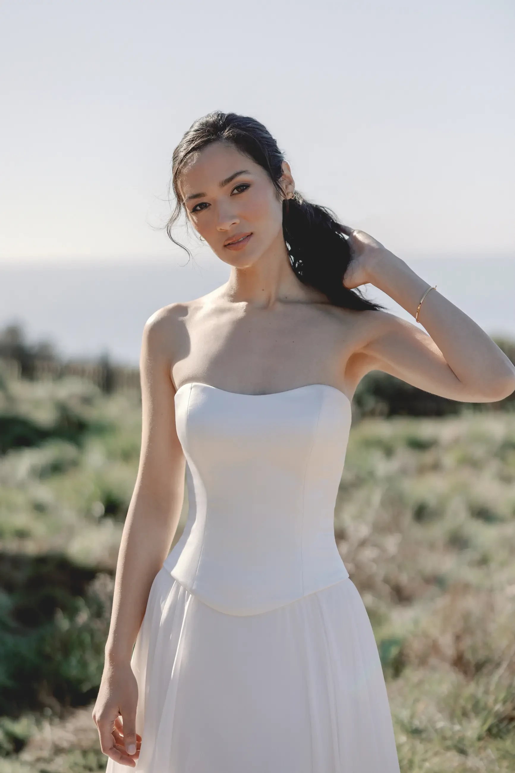 A woman in a strapless white dress stands outdoors, gently holding her hair. The scene is serene with a blurred grassy background and a soft, natural light.