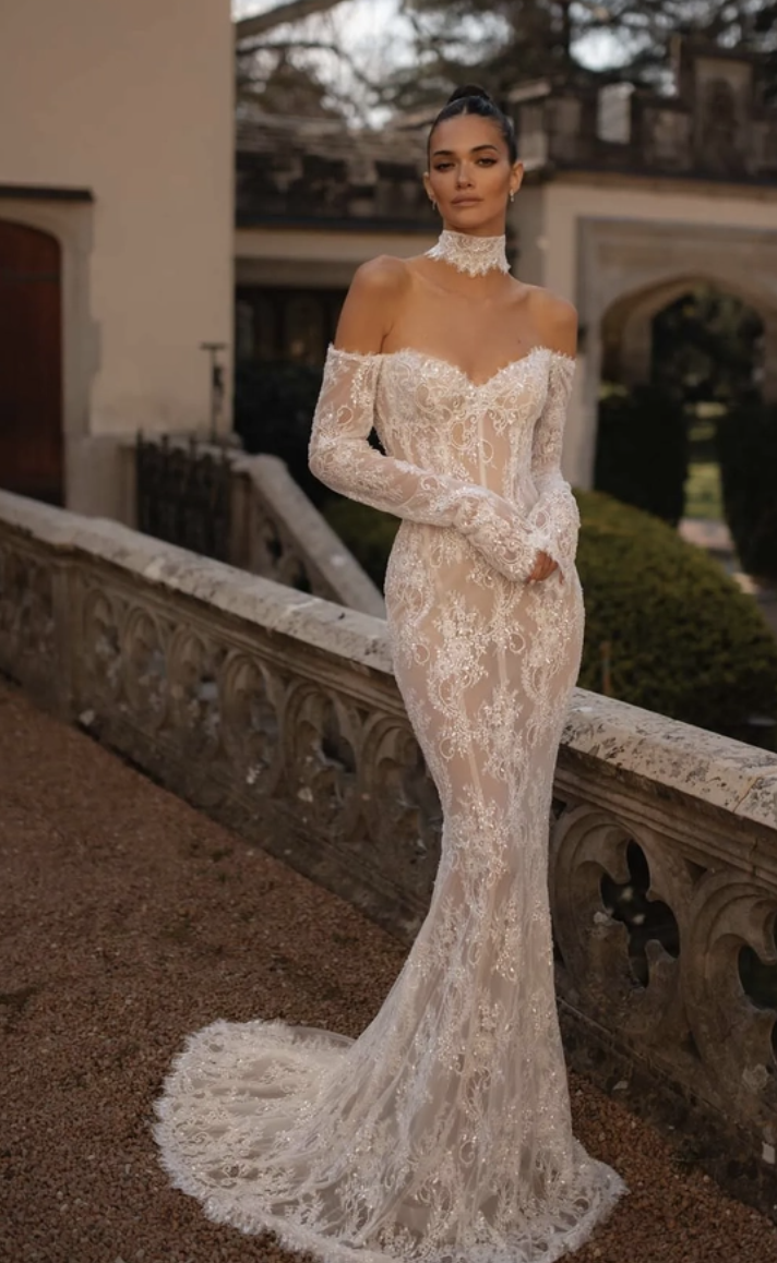 A model wearing an elegant lace wedding gown with off-the-shoulder sleeves, posing outside in a scenic setting.