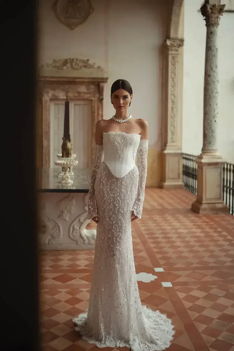 A woman in a fitted, long-sleeve white gown with intricate lace details, standing elegantly in an ornate indoor setting with historical architecture.