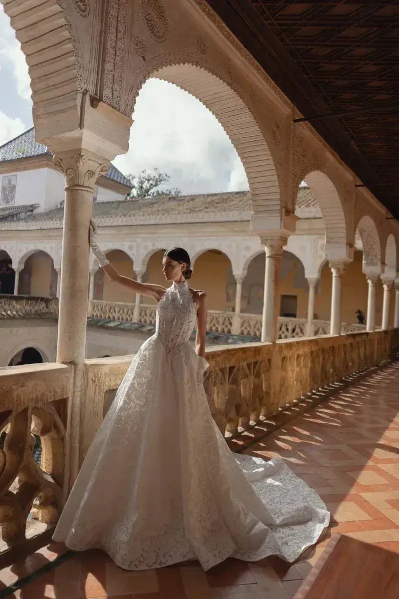 A bride in a flowing white gown stands gracefully near an ornate archway, holding a fan, with sunlight casting shadows across the intricately tiled floor and architecture.