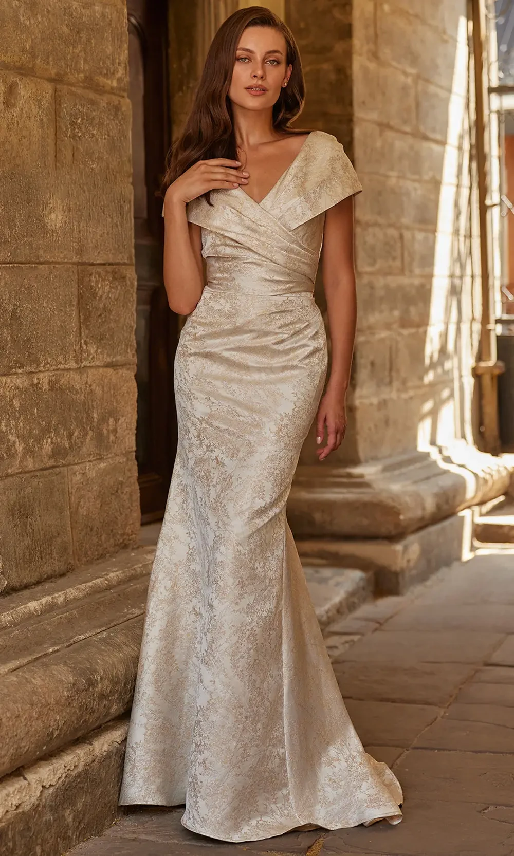 A woman standing elegantly in a form-fitting, off-white gown with a v-neckline and cap sleeves, against a backdrop of stone architecture.