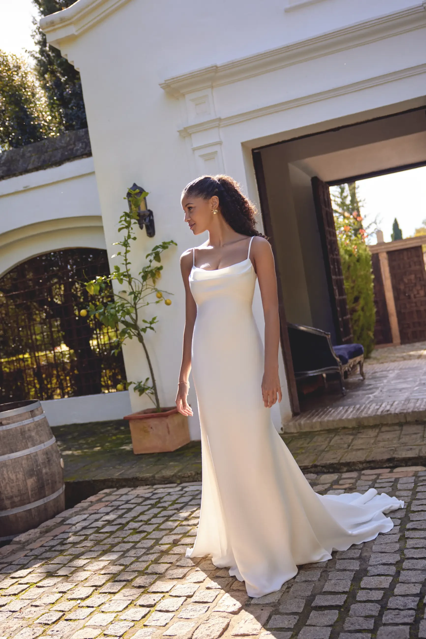 A woman in an elegant white wedding gown stands outdoors on a cobbled path. She is near a white building with a lantern and potted plant, exuding a serene and sophisticated mood.