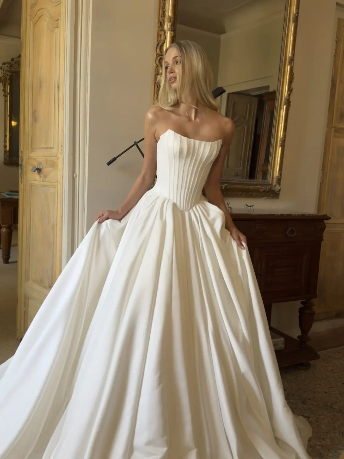 A woman in a strapless, floor-length white gown poses elegantly in a well-decorated room with wooden furniture and ornate mirrors.