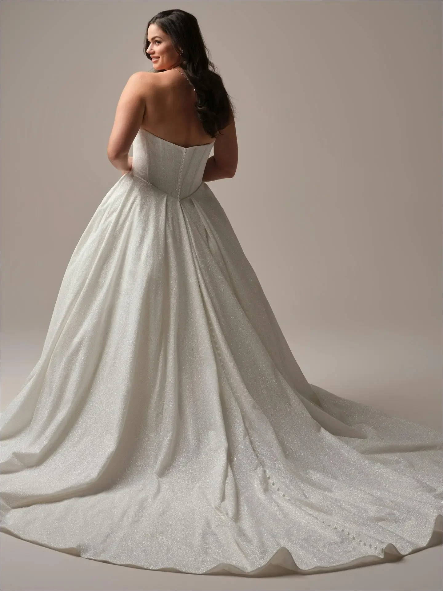 Woman in an elegant strapless wedding gown stands with her back to the camera, long wavy hair flowing over her shoulder. The dress has a sweeping train.