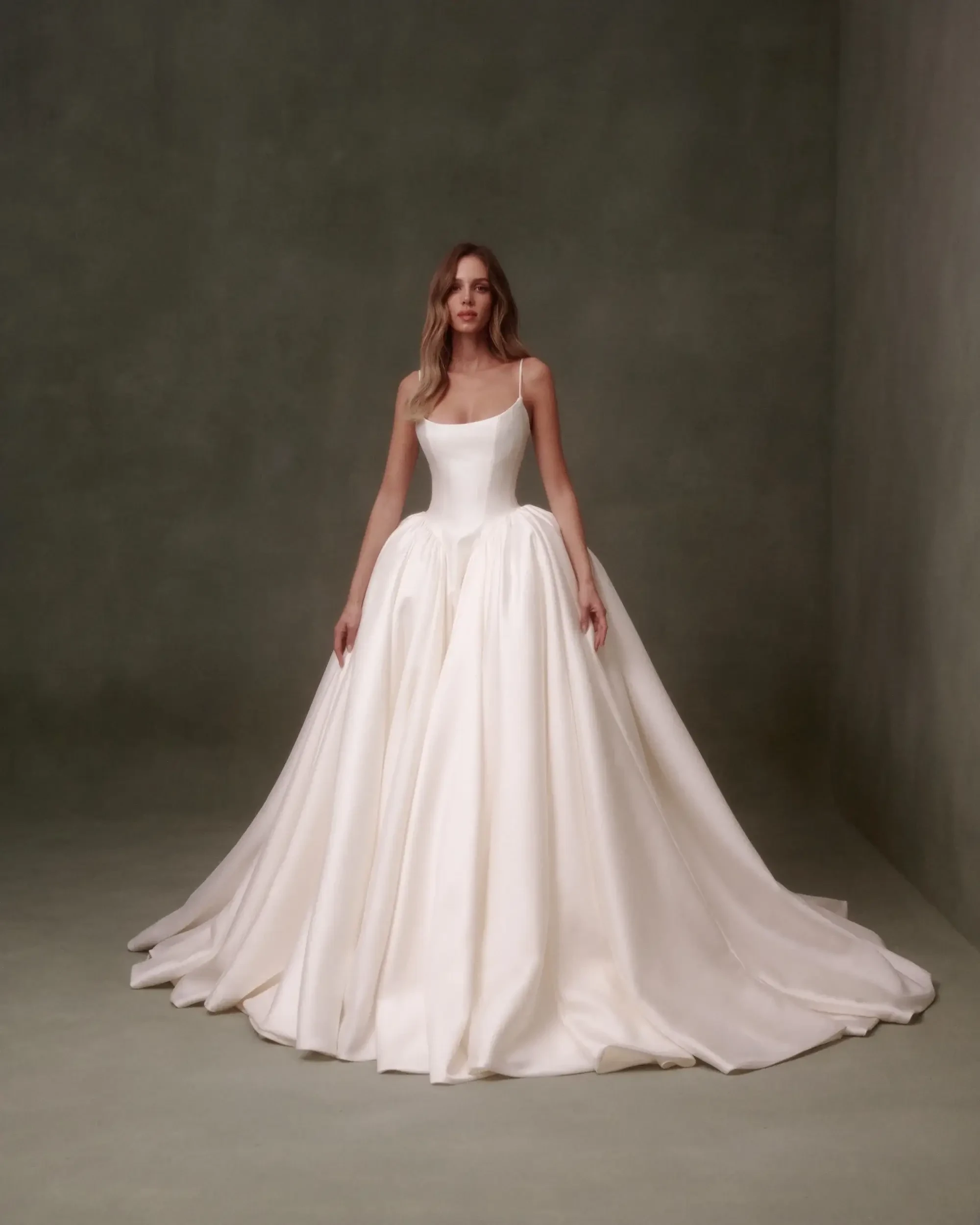 A woman wearing a large, flowing white wedding dress stands against a minimalistic background.