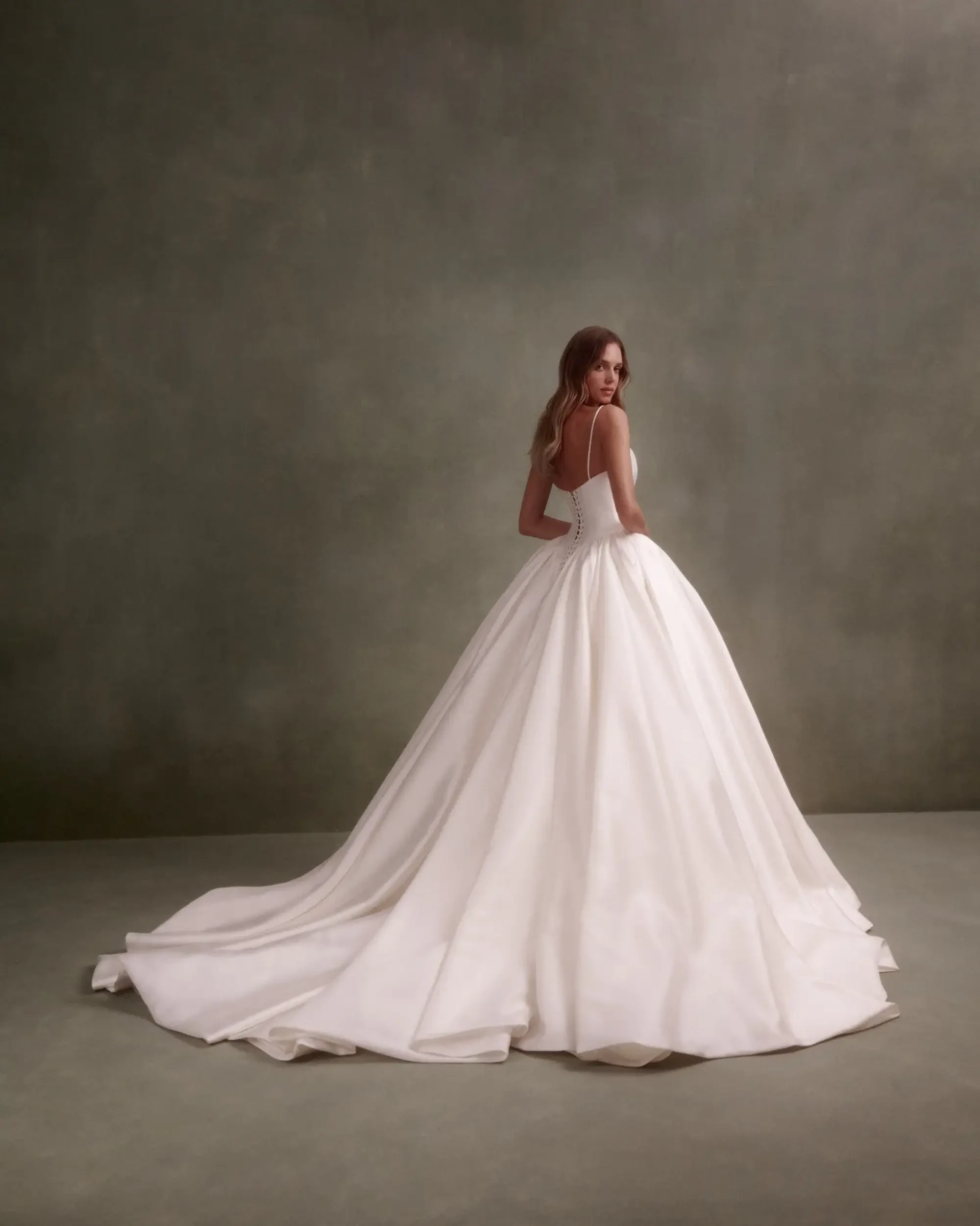 A woman in a beautiful white gown stands with her back to the camera, showcasing a long train in a neutral, softly lit setting.