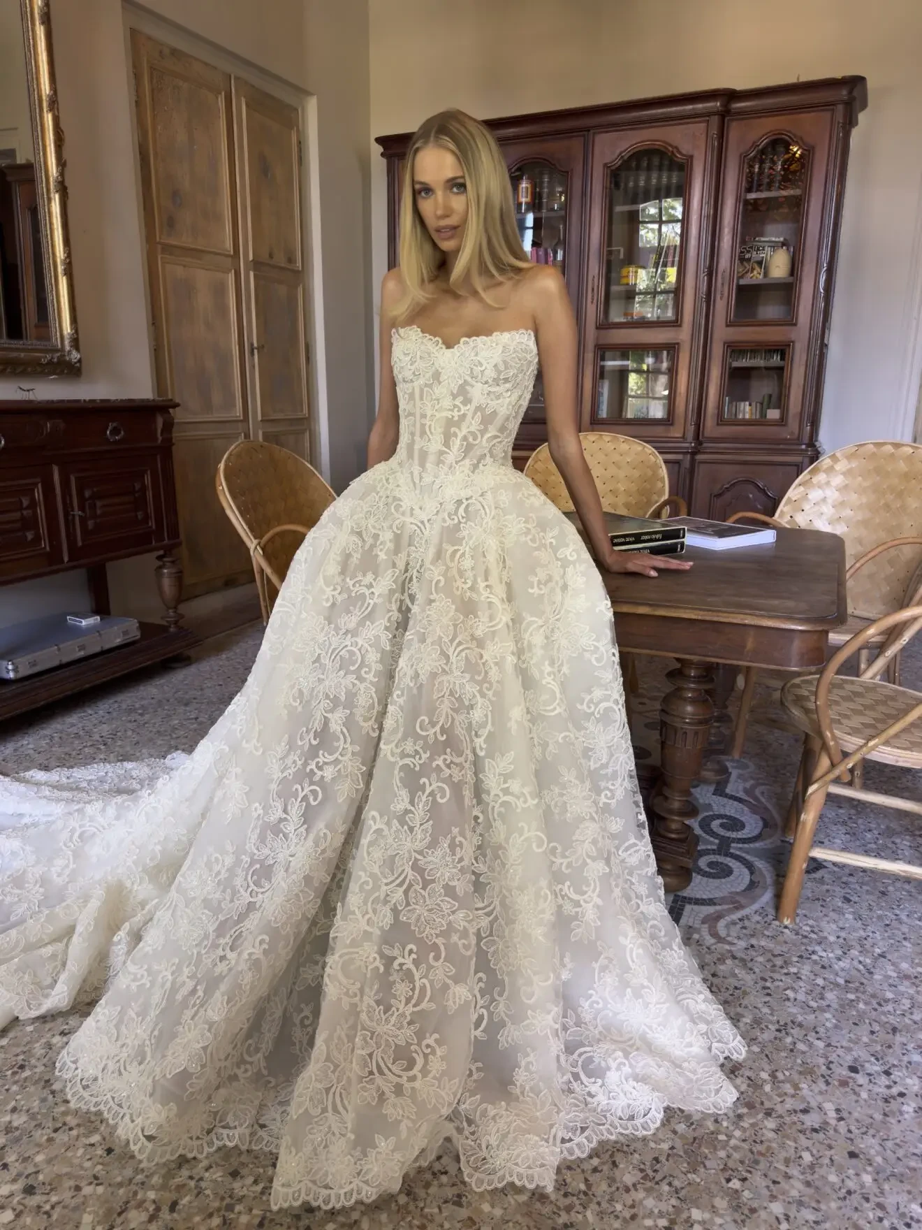 A woman in a stunning lace wedding gown stands in a well-lit room with a wooden table and chairs, surrounded by cabinets.