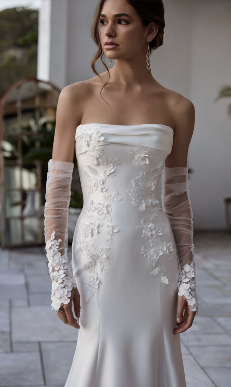 A model wearing a strapless white wedding gown with floral embellishments and sheer sleeves.