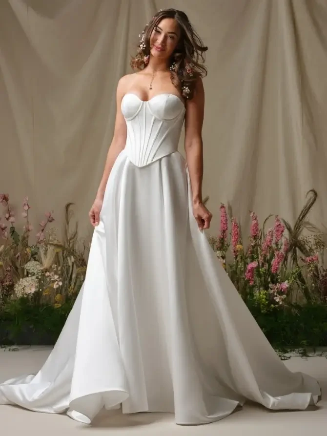 A woman in a strapless, white wedding gown with a corset bodice standing against a soft beige backdrop with floral arrangements.