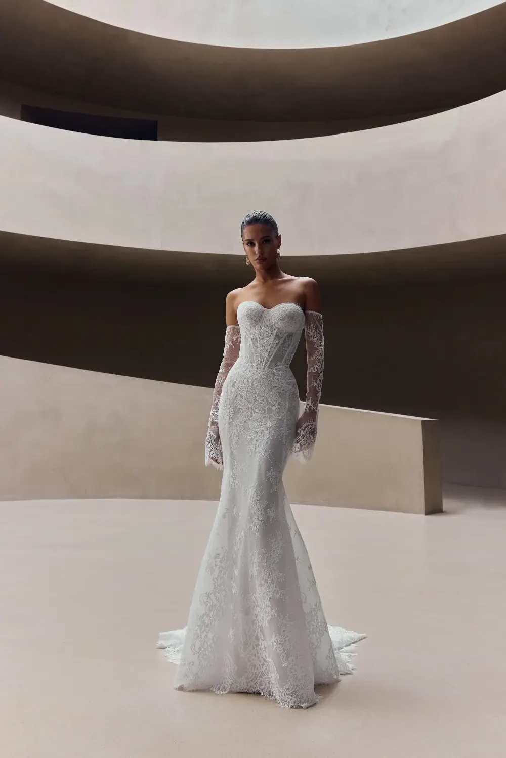A model wearing a strapless white lace wedding gown with long gloves, standing in an elegantly minimalist space.