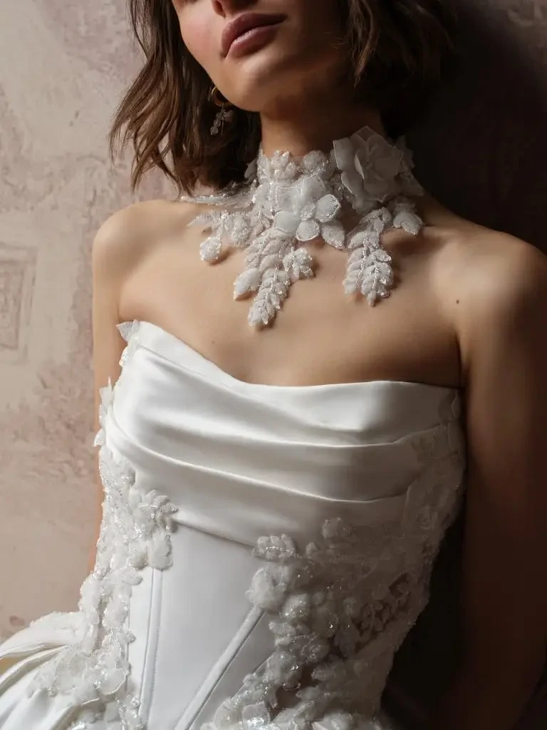 A model wearing an elegant white ball gown with intricate floral embroidery and a strapless neckline, posing in a softly lit, artistic background resembling a classic painting.