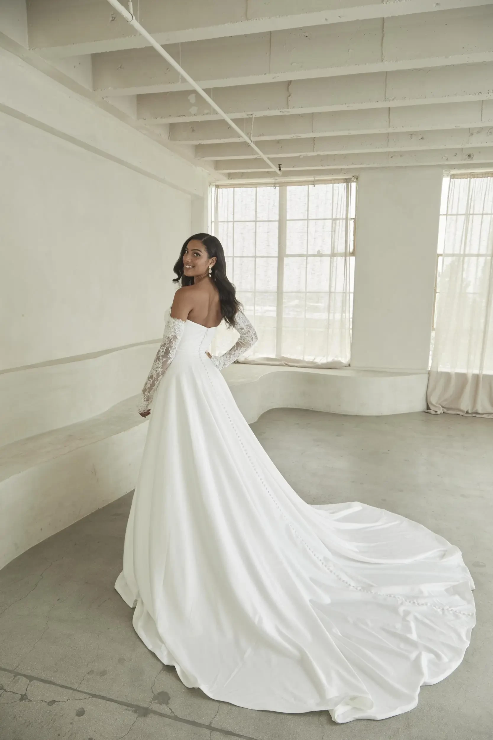 A woman in an elegant off-the-shoulder wedding dress with lace sleeves stands in a bright, minimalist room. Her gown's long train cascades gracefully.