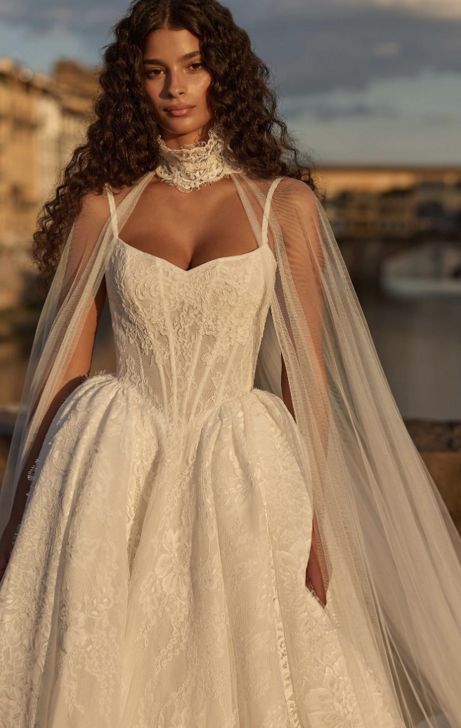 A model in a white wedding dress with a fitted bodice, flowing skirt, and sheer cape-like sleeves, standing outdoors with a picturesque background.
