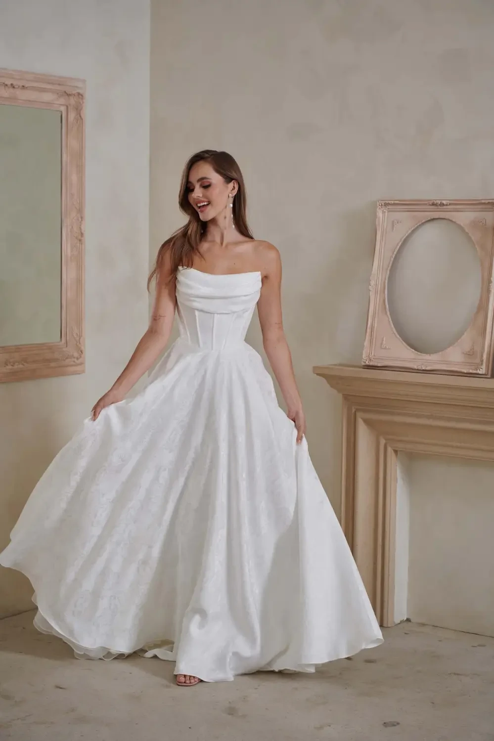 A woman in a white strapless gown standing gracefully in a room with neutral-colored walls and a decorative mirror.
