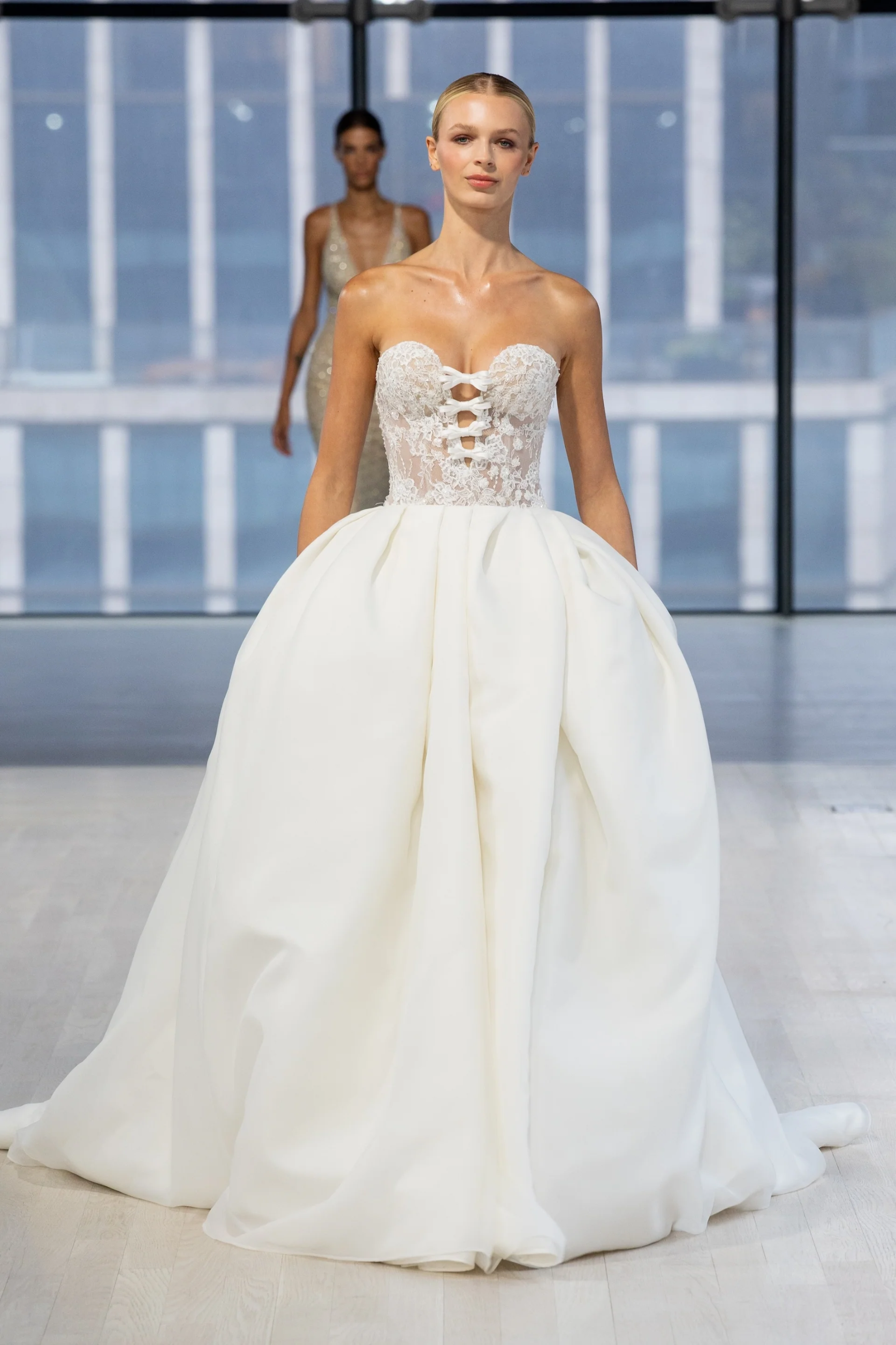 A model in a strapless wedding dress with a fitted lace bodice and a full, flowing skirt walks down the runway. Another model in a shimmering gown is visible in the background.