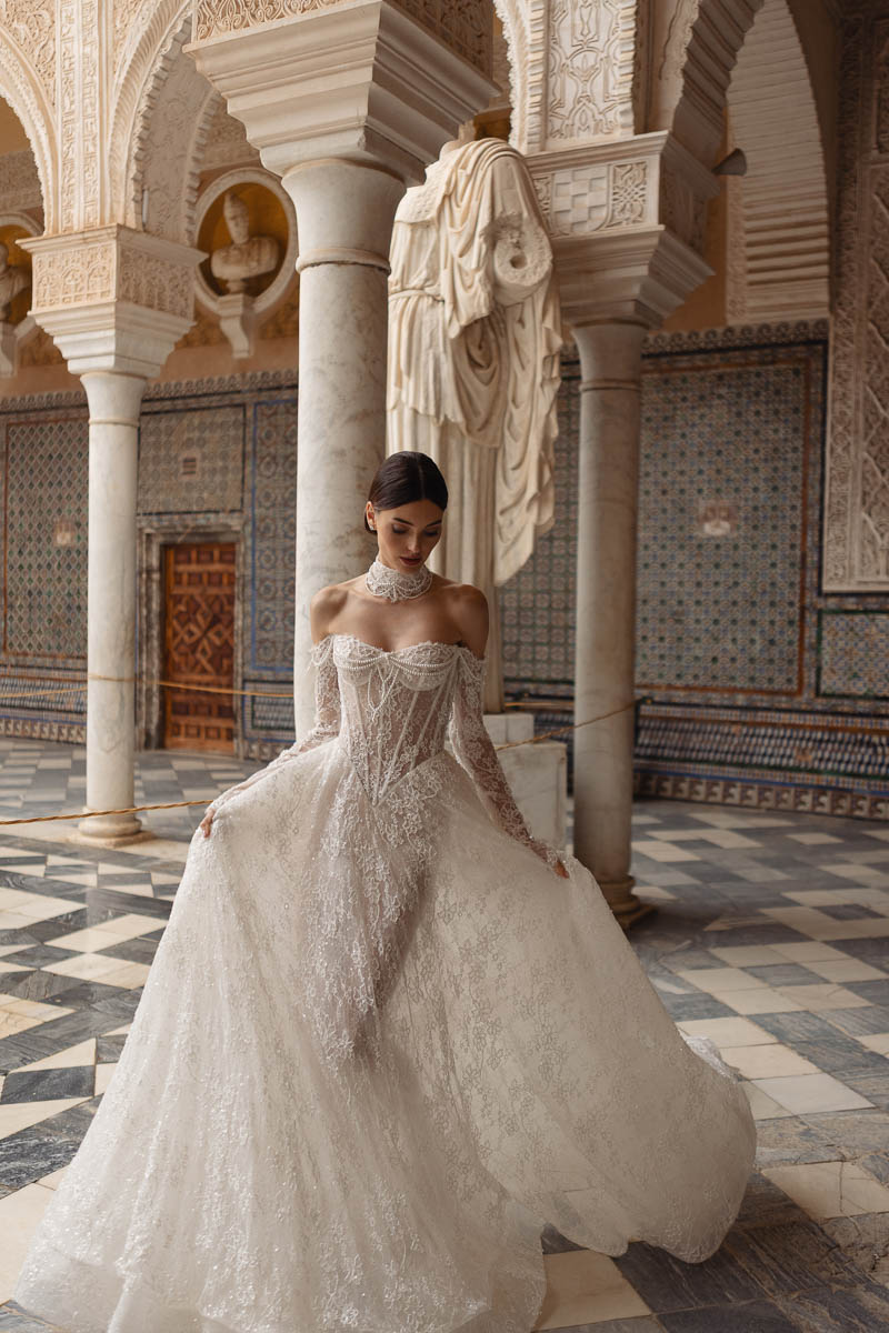 A woman wearing an elaborate white wedding dress stands gracefully in a beautifully decorated space with intricate architectural details.