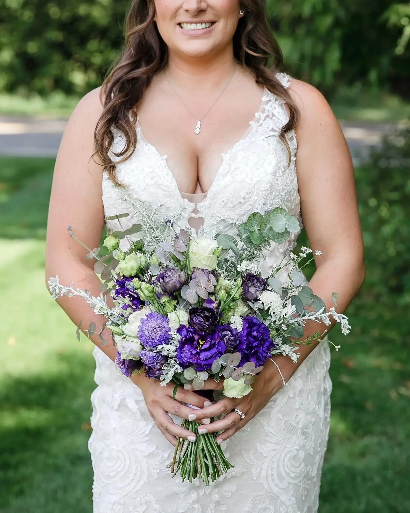 A smiling woman in a lace wedding dress holding a bouquet of purple and white flowers.