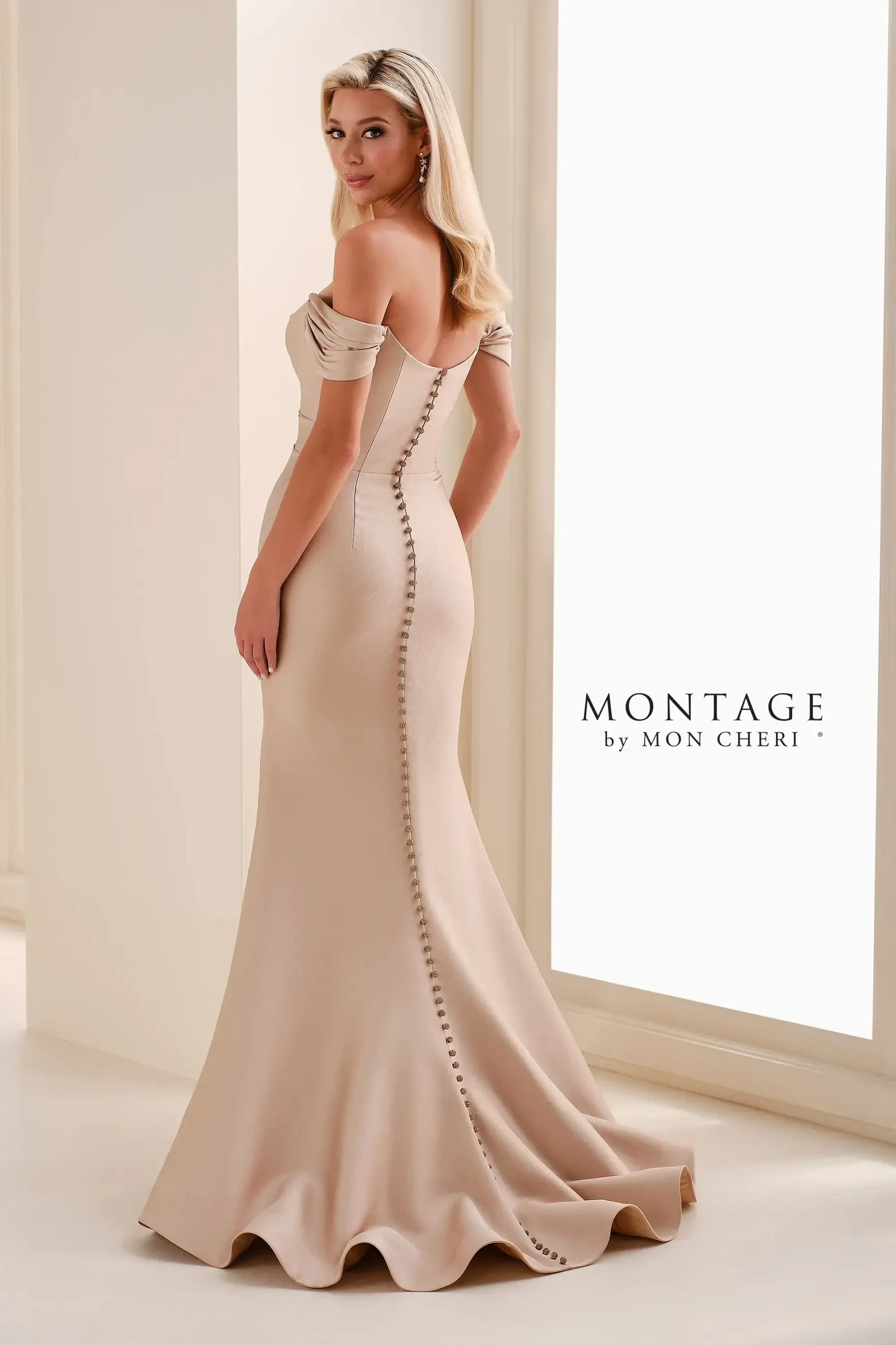 A woman in an elegant, off-the-shoulder beige gown with a fitted silhouette and buttons down the back, standing gracefully in front of a softly lit backdrop.