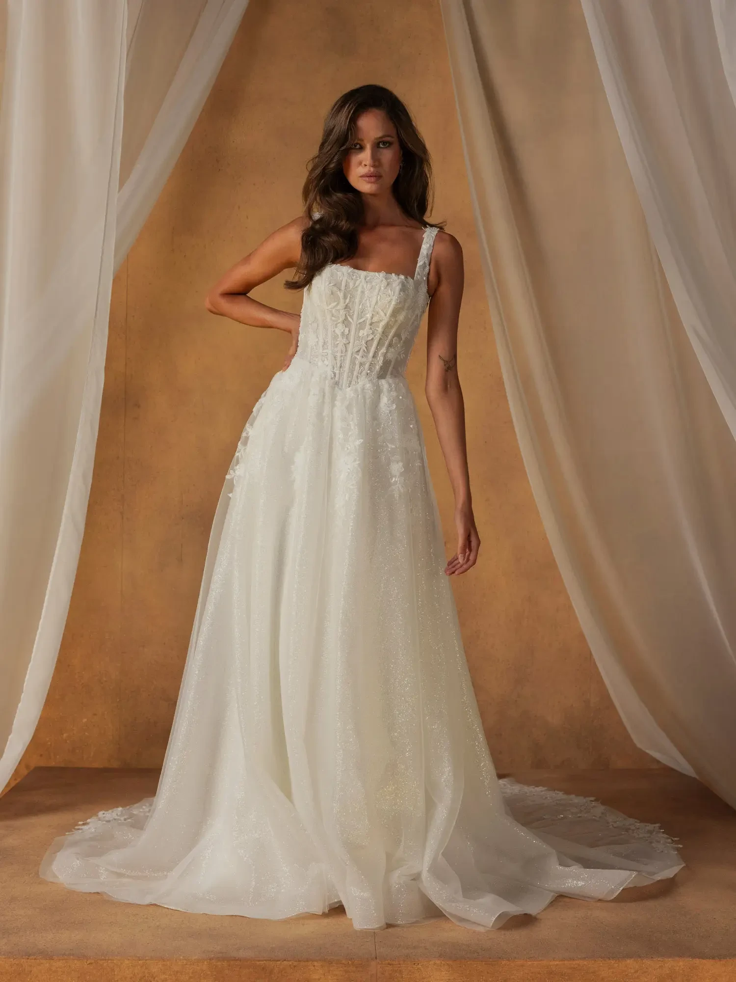 A woman stands in an elegant, flowing white wedding gown with lace details. The backdrop features soft, beige drapes, conveying a romantic ambiance.