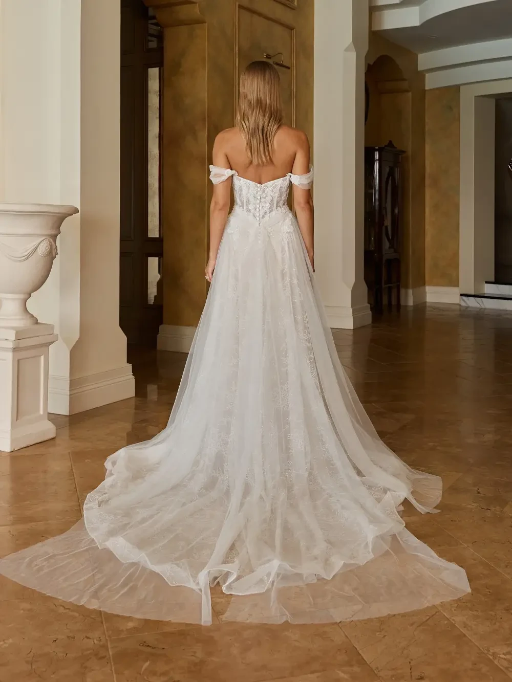 A woman in an elegant, off-the-shoulder white wedding dress standing in an indoor setting with warm-colored walls and a tiled floor.