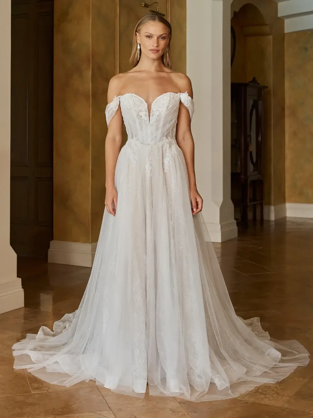 A woman in an elegant, off-the-shoulder white wedding dress standing in an indoor setting with warm-colored walls and a tiled floor.