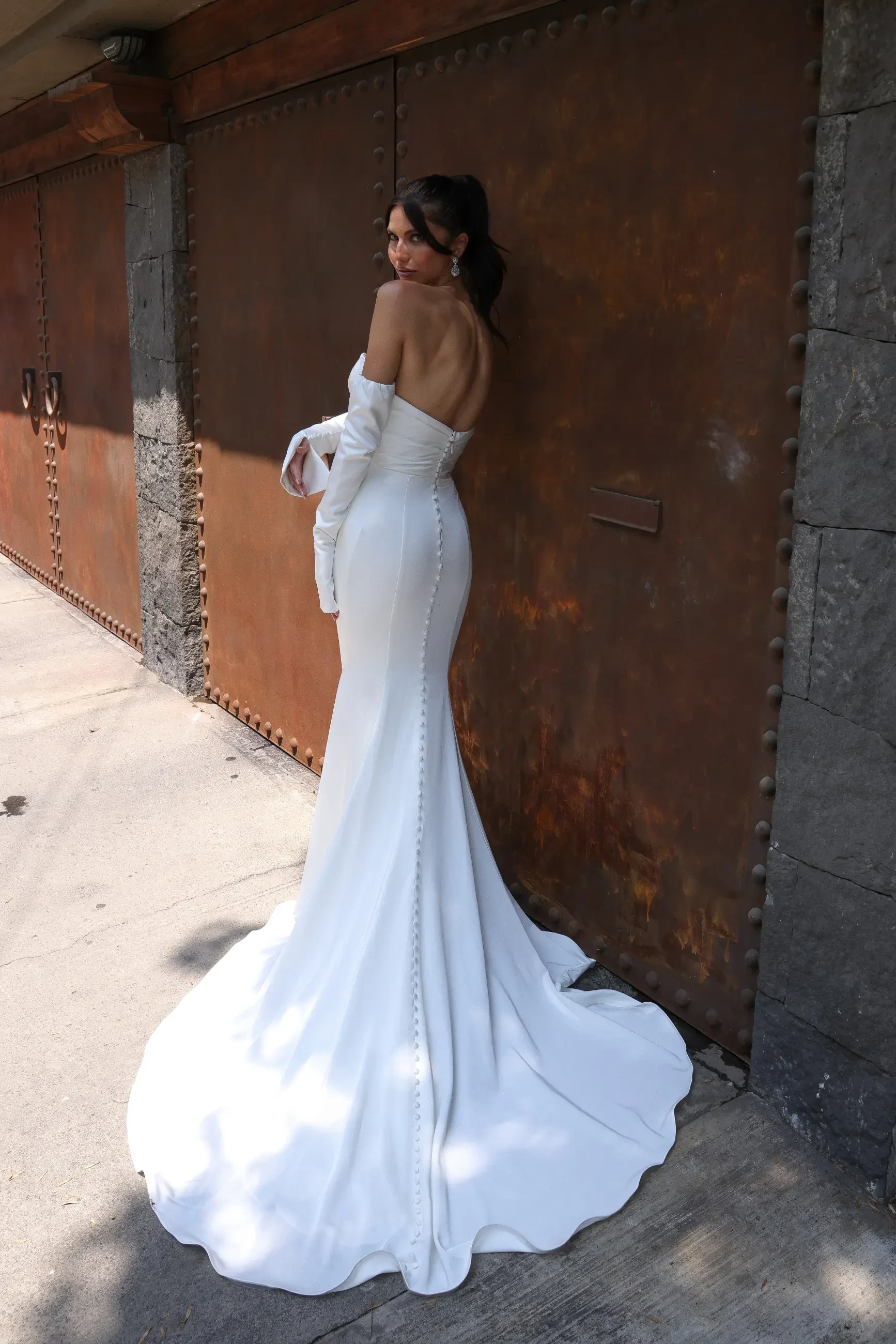 A woman in an elegant, strapless white gown with long gloves stands against a rustic metal door. The mood is sophisticated and classic.