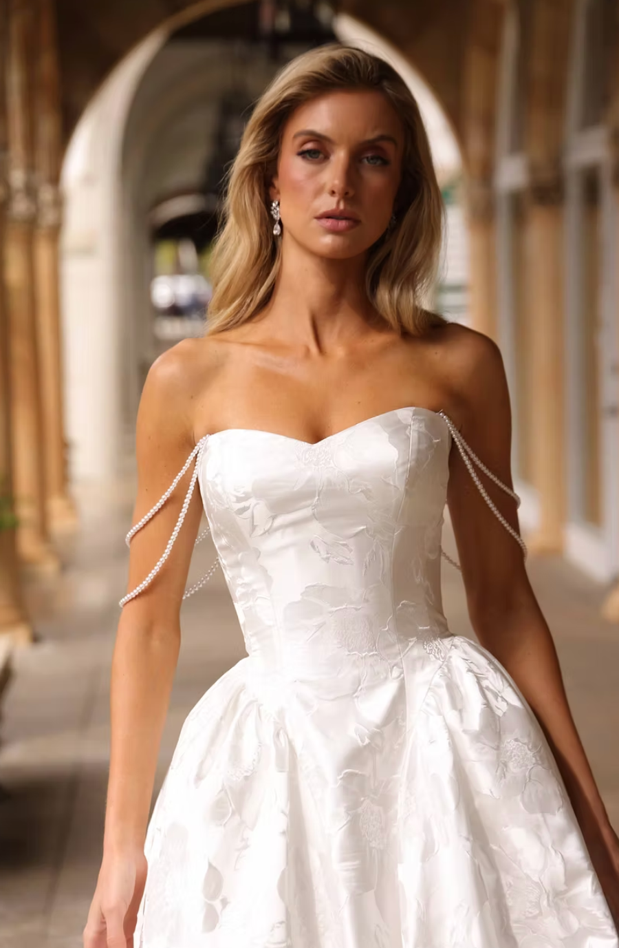 A woman in a strapless white wedding dress with pearl detailing, standing in an arched corridor.